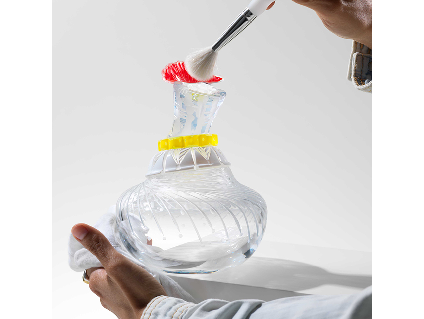 a photograph of a glass bottle. It is held by a hand at its base, and is being dusted by another hand at the top. The bottle is transparent clear glass, with a yellow detail at the top and a red stopper.