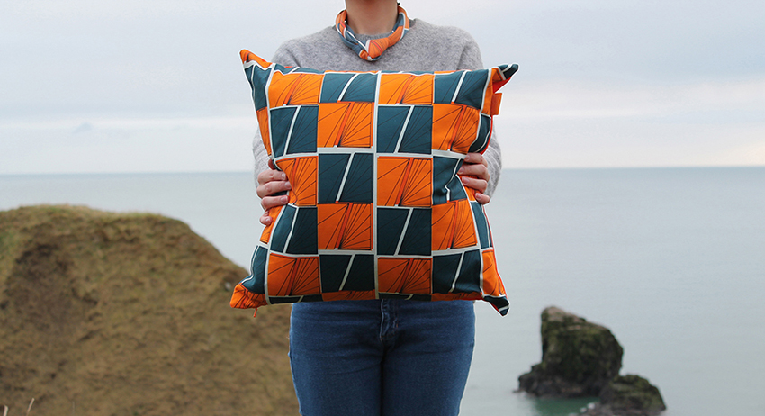 Model wearing patterned scarf holds patterned cushion in front of the sea